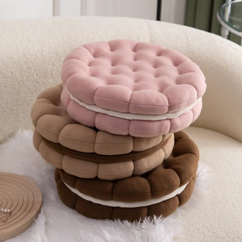 Stack of biscuit shaped plush cushions in strawberry pink, chocolate coffee, and almond beige on a white sofa.