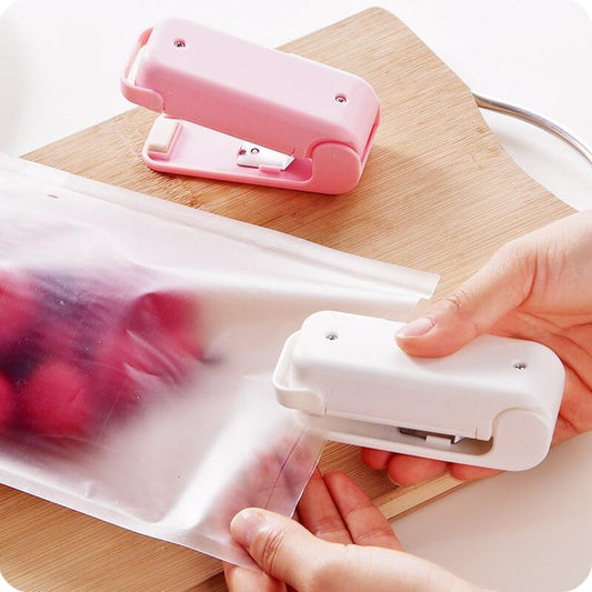 Pink and white Mini heat bag sealer device creating airtight seal on snack bag to preserve food freshness on a wooden surface with hands using one of them.