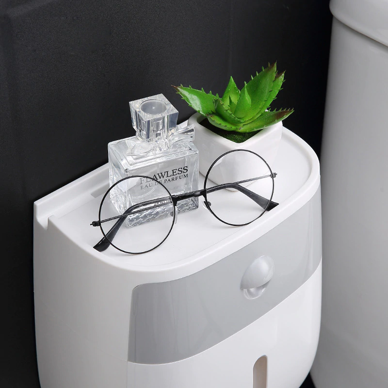 Small white wall mounted waterproof toilet paper holder with storage drawer with a plant, glasses, and perfume bottle against a black background