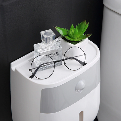 Small white wall mounted waterproof toilet paper holder with storage drawer with a plant, glasses, and perfume bottle against a black background